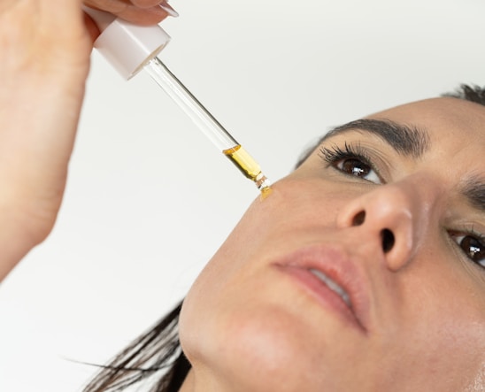 A close-up of a person applying a skincare serum or oil to their cheek using a dropper. The person has neutral expression with a focus on skincare and wellness.