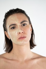 A person with shoulder-length hair is shown up close against a neutral background. Half of their face is covered in a translucent facial mask or gel, giving the skin a glossy appearance. The pose is calm and direct, with a neutral expression.
