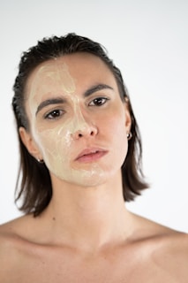 A person with shoulder-length hair is shown up close against a neutral background. Half of their face is covered in a translucent facial mask or gel, giving the skin a glossy appearance. The pose is calm and direct, with a neutral expression.