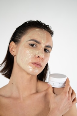 A person with short dark hair applies a transparent facial mask or cream to their face, holding a jar of the product in one hand against a plain, light background.