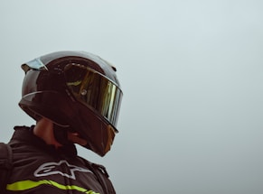 A person wearing a sleek, dark motorcycle helmet with a reflective visor is gazing sideways. The helmet has droplets of water on its surface. The background is a plain, light gray sky.