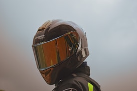A person is wearing a glossy motorcycle helmet with a reflective visor that shows potential reflections of the surroundings. The helmet has a branded name on the top and appears to be part of riding gear. The background is a muted, overcast sky.