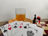 A hand of playing cards featuring four aces and a three of clubs is displayed over a checkered board. A glass of amber liquid rests on an orange saucer in the background. There are stacked poker chips, dice, and a black chess piece nearby.