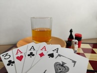 A hand of playing cards featuring four aces and a three of clubs is displayed over a checkered board. A glass of amber liquid rests on an orange saucer in the background. There are stacked poker chips, dice, and a black chess piece nearby.