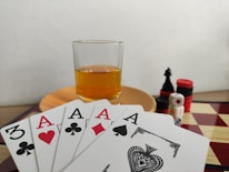 A hand of playing cards featuring four aces and a three of clubs is displayed over a checkered board. A glass of amber liquid rests on an orange saucer in the background. There are stacked poker chips, dice, and a black chess piece nearby.