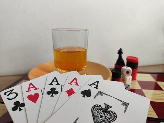 A hand of playing cards featuring four aces and a three of clubs is displayed over a checkered board. A glass of amber liquid rests on an orange saucer in the background. There are stacked poker chips, dice, and a black chess piece nearby.