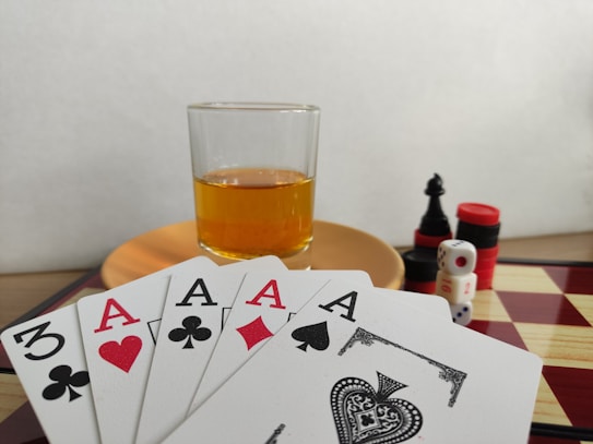 A hand of playing cards featuring four aces and a three of clubs is displayed over a checkered board. A glass of amber liquid rests on an orange saucer in the background. There are stacked poker chips, dice, and a black chess piece nearby.