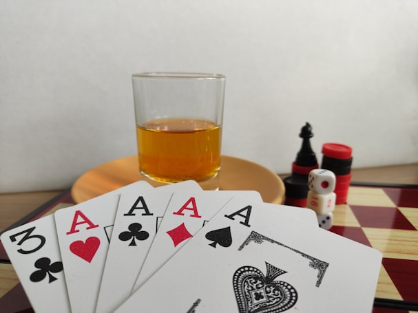 A hand of playing cards featuring four aces and a three of clubs is displayed over a checkered board. A glass of amber liquid rests on an orange saucer in the background. There are stacked poker chips, dice, and a black chess piece nearby.