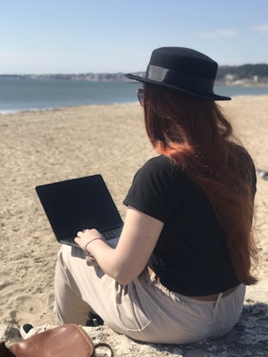 A person with long red hair is sitting on a sandy beach, working on a laptop. They are wearing a black hat and sunglasses, facing the ocean under a clear blue sky.