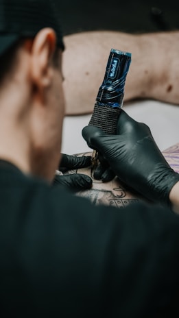 A person wearing black gloves is holding a tattoo machine, applying ink on someone's arm. The arm is resting on a padded surface, and the tattoo design includes geometric patterns and lines.