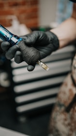 A person wearing black gloves holds a tattoo machine. The machine has a black grip and blue accents, set against a blurred background of a workshop with drawers and tools.