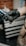 A person wearing black gloves holds a tattoo machine. The machine has a black grip and blue accents, set against a blurred background of a workshop with drawers and tools.