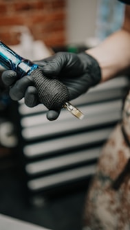 A person wearing black gloves holds a tattoo machine. The machine has a black grip and blue accents, set against a blurred background of a workshop with drawers and tools.
