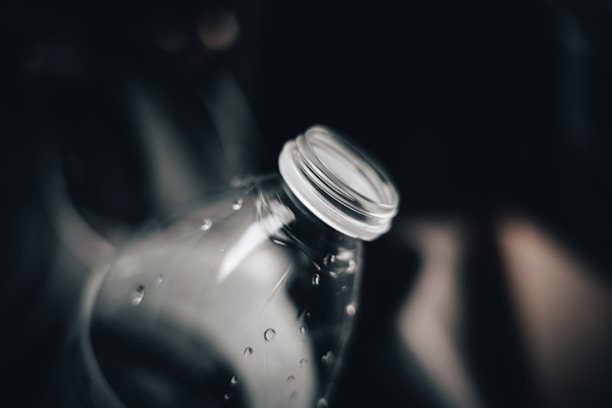 A close-up of an empty, transparent plastic bottle with a focus on the neck and cap area. Water droplets are visible on the surface of the bottle, and the background is dark and blurred, creating a moody and dramatic effect.