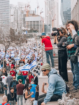 A large crowd of people is gathered in an urban area, many carrying flags with a blue and white design. Several individuals stand on an elevated platform, some are taking photos with professional cameras. The background features tall modern buildings and construction cranes.