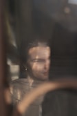 A softly focused reflection of a person with long hair is visible through a glass surface. They appear to be partially obscured by shadows, with subtle details of their clothing and facial features blurred. The ambient light creates a dreamy and introspective atmosphere.