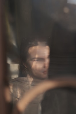 A softly focused reflection of a person with long hair is visible through a glass surface. They appear to be partially obscured by shadows, with subtle details of their clothing and facial features blurred. The ambient light creates a dreamy and introspective atmosphere.