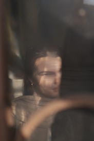 A softly focused reflection of a person with long hair is visible through a glass surface. They appear to be partially obscured by shadows, with subtle details of their clothing and facial features blurred. The ambient light creates a dreamy and introspective atmosphere.