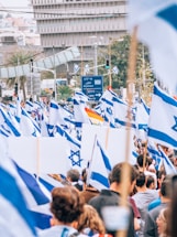 A large crowd is gathered for a demonstration or rally, holding numerous Israeli flags. The scene is bustling with people, and a large road sign in Hebrew is visible in the background, situated in an urban area.