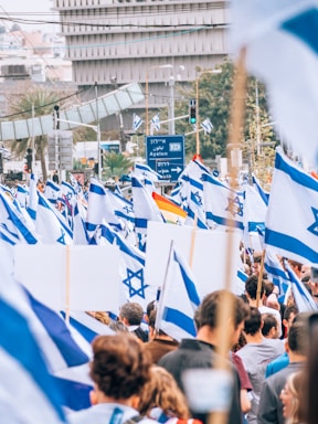 A large crowd is gathered for a demonstration or rally, holding numerous Israeli flags. The scene is bustling with people, and a large road sign in Hebrew is visible in the background, situated in an urban area.
