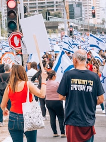 A crowd of people is gathered for a protest or demonstration. Many individuals are holding Israeli flags, while a woman in a red top is holding a blank protest sign. The scene is busy, with a variety of people of different ages present. Traffic lights and a stop sign are visible, indicating the event is taking place at a street intersection in an urban area. A person wearing a shirt with text in another language is also in the foreground.