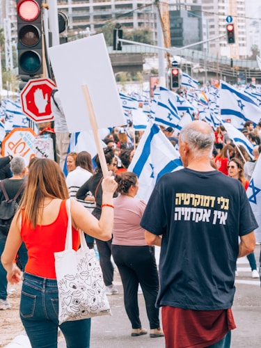 A crowd of people is gathered for a protest or demonstration. Many individuals are holding Israeli flags, while a woman in a red top is holding a blank protest sign. The scene is busy, with a variety of people of different ages present. Traffic lights and a stop sign are visible, indicating the event is taking place at a street intersection in an urban area. A person wearing a shirt with text in another language is also in the foreground.