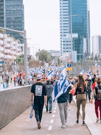 A large group of people is walking along a city street holding Israeli flags. The scene is set in an urban area with tall modern buildings lining the street. Some individuals wear casual clothing, while others hold signs, possibly indicating a protest or march.