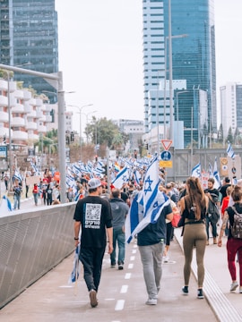 A large group of people is walking along a city street holding Israeli flags. The scene is set in an urban area with tall modern buildings lining the street. Some individuals wear casual clothing, while others hold signs, possibly indicating a protest or march.