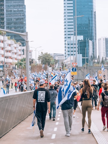 A large group of people is walking along a city street holding Israeli flags. The scene is set in an urban area with tall modern buildings lining the street. Some individuals wear casual clothing, while others hold signs, possibly indicating a protest or march.