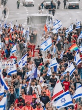 A large group of people are participating in a protest or demonstration on a street, carrying numerous Israeli flags. Some participants hold rainbow flags, and there are various signs visible, suggesting diverse messages and beliefs. Vehicles and security personnel are present in the background, indicating a controlled environment. The crowd appears dense and engaged in the activity.