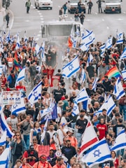 A large group of people are participating in a protest or demonstration on a street, carrying numerous Israeli flags. Some participants hold rainbow flags, and there are various signs visible, suggesting diverse messages and beliefs. Vehicles and security personnel are present in the background, indicating a controlled environment. The crowd appears dense and engaged in the activity.