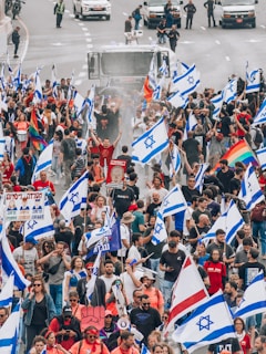 A large group of people are participating in a protest or demonstration on a street, carrying numerous Israeli flags. Some participants hold rainbow flags, and there are various signs visible, suggesting diverse messages and beliefs. Vehicles and security personnel are present in the background, indicating a controlled environment. The crowd appears dense and engaged in the activity.
