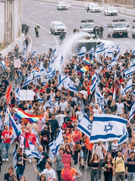 A large group of people are gathered in a protest or demonstration on a highway. Many individuals are holding flags with a blue Star of David, and some carry rainbow flags. The scene has vehicles, including police cars and a water cannon, creating a dynamic and tense atmosphere.