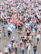 A large group of people are gathered on a street, participating in a demonstration or march. Many are holding flags with a blue Star of David on a white background. Some individuals are also carrying rainbow flags, suggesting support for LGBTQ+ rights. The crowd is diverse, with people of various ages and styles. The atmosphere seems vibrant and energetic.