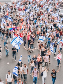 A large group of people are gathered on a street, participating in a demonstration or march. Many are holding flags with a blue Star of David on a white background. Some individuals are also carrying rainbow flags, suggesting support for LGBTQ+ rights. The crowd is diverse, with people of various ages and styles. The atmosphere seems vibrant and energetic.