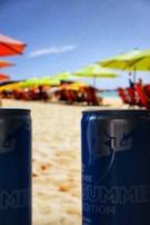 Cans of a summer edition drink prominently displayed in the foreground on a sandy beach, with colorful beach umbrellas and chairs visible in the blurred background. The scene is under a clear blue sky.