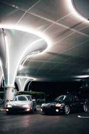 Two cars are parked under a modern, illuminated canopy with a sleek architectural design. The setting is nighttime, creating a dramatic contrast between the glowing white structure above and the dark surroundings. A bus is visible in the background, adding context to the location as a possible transportation hub.