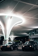 A modern, well-lit parking area features two dark cars parked under uniquely designed, large white structures that illuminate the scene. The area includes curved architecture with windows and signage indicating a commercial or public space. The lighting provides a futuristic and sleek ambiance.