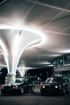A modern, well-lit parking area features two dark cars parked under uniquely designed, large white structures that illuminate the scene. The area includes curved architecture with windows and signage indicating a commercial or public space. The lighting provides a futuristic and sleek ambiance.