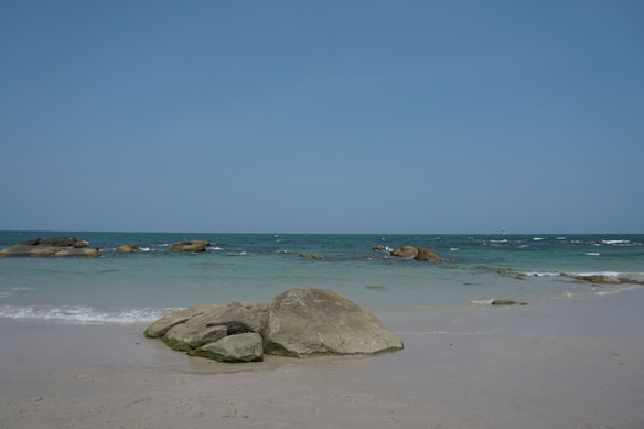 A tranquil beach scene with clear blue skies and gentle waves lapping against large rocks on a sandy shore. The water is a mix of turquoise and deep blue, creating a sense of calm and openness.