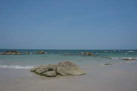 A tranquil beach scene with clear blue skies and gentle waves lapping against large rocks on a sandy shore. The water is a mix of turquoise and deep blue, creating a sense of calm and openness.