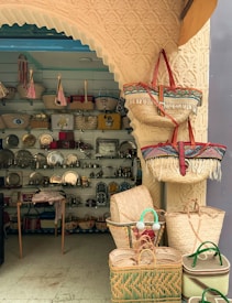 A quaint shop entrance displaying a variety of woven baskets, some hanging and some stacked. Inside, shelves are filled with metallic decorative items and colorful tote bags. The arched entrance features intricate patterns.