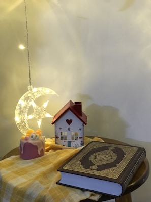 A cozy tabletop display includes a small decorative house with a red roof, a bright moon and star lamp, a pink decorative candle with small fruits, and a book with ornate gold detailing on the cover. The table is covered with a yellow checkered cloth, and the background wall is softly lit, creating a warm atmosphere.