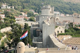 A picturesque stone fortress wall winds through a historic town with lush greenery in the background. The setting appears to be on a hillside with numerous small, charming houses with red-tiled roofs. A Croatian flag waves prominently from a tower, as visitors walk along the fortified walls.
