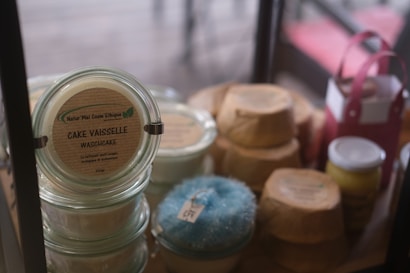 Various jars and recyclable packaging are arranged neatly, featuring a product labeled 'Cake Vaisselle Waschcake'. These are eco-friendly cleaning products displayed on a shelf, with a focus on sustainability and aesthetics. A pink container and a yellow jar are visible among the items.