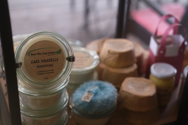 Various jars and recyclable packaging are arranged neatly, featuring a product labeled 'Cake Vaisselle Waschcake'. These are eco-friendly cleaning products displayed on a shelf, with a focus on sustainability and aesthetics. A pink container and a yellow jar are visible among the items.