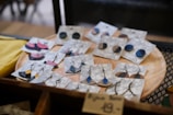A collection of handmade earrings is displayed on marble-patterned cards, resting on a wooden circular tray. The earrings come in various shapes and colors, including blue, black, and pink, with both stud and dangling designs featuring metal chains and hooks. The display gives a rustic and artisanal feel.