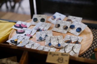 A collection of handmade earrings is displayed on marble-patterned cards, resting on a wooden circular tray. The earrings come in various shapes and colors, including blue, black, and pink, with both stud and dangling designs featuring metal chains and hooks. The display gives a rustic and artisanal feel.