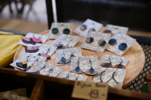 A collection of handmade earrings is displayed on marble-patterned cards, resting on a wooden circular tray. The earrings come in various shapes and colors, including blue, black, and pink, with both stud and dangling designs featuring metal chains and hooks. The display gives a rustic and artisanal feel.