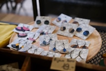 A collection of handmade earrings is displayed on marble-patterned cards, resting on a wooden circular tray. The earrings come in various shapes and colors, including blue, black, and pink, with both stud and dangling designs featuring metal chains and hooks. The display gives a rustic and artisanal feel.